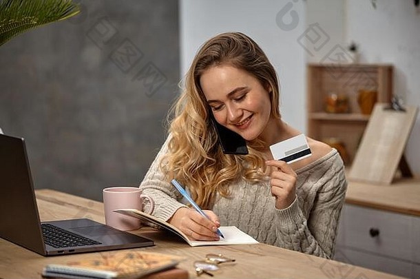 穿着米色毛衣的女博主。微笑着，坐在厨房的桌子旁，用智能手机聊天，在笔记本上写点东西，展示塑料卡片