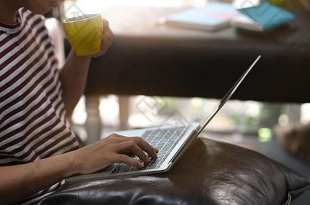一张裁剪过的图片，创意人一边喝咖啡，一边坐在皮革店放松，一边在膝上的笔记本电脑上使用/打字