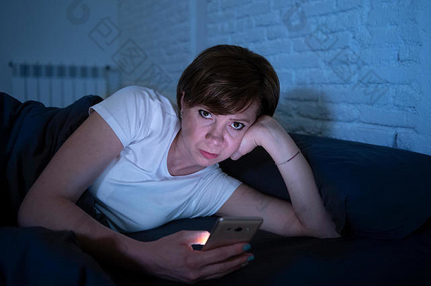 一位年轻迷人的女士躺在黑暗的卧室里，深夜用智能手机醒来。使用手机进行网上聊天和发送信息