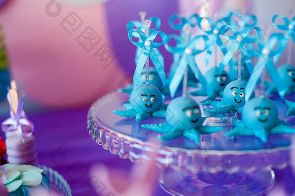 蓝色的蛋糕上有有趣的章鱼，放在玻璃圆盘子上，还有紫色背景上有棉花糖的罐子。生日派对上的夏日糖果吧