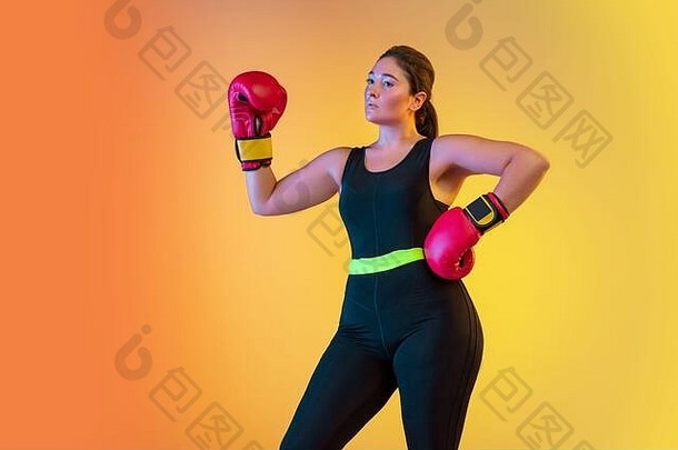 年轻的白人大个子女模特在霓虹灯下的渐变橙色桃色背景上训练。戴拳击手套训练。体育观念、健康生活方式、身体积极、平等。