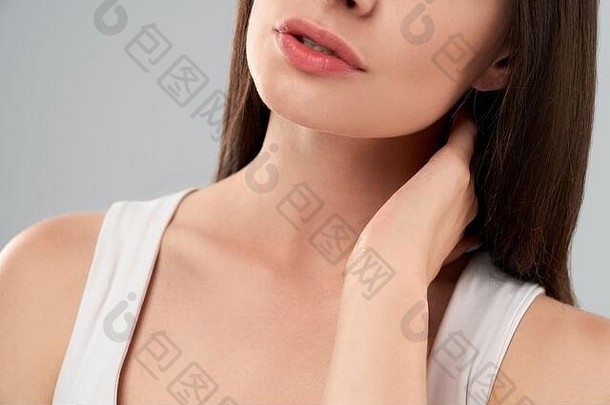 一名穿着白衬衫的不知名白人妇女在灰色隔离背景上摆姿势，因疼痛用手触摸脖子。前茬黑发用嘴呼吸。
