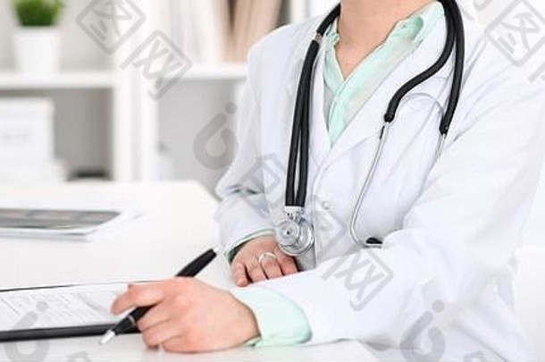 在医院办公室的工作场所，一名女医生坐在办公桌旁，手里拿着电脑。无<strong>名医</strong>生的手特写。衬衫的绿调。医药