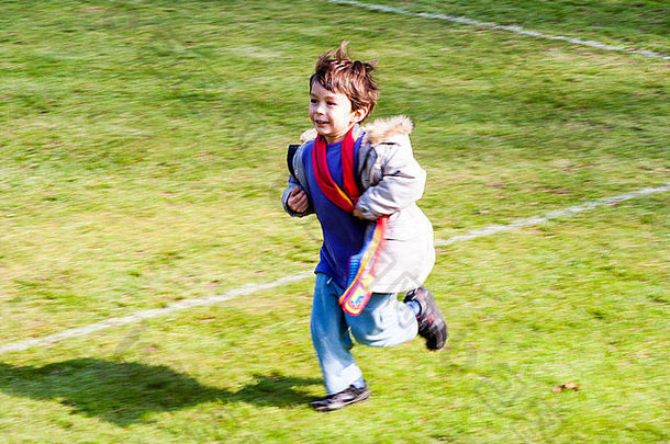 高加索儿童，男孩，5-6岁，穿着外套，戴着围巾，沿着画着白线的运动草坪快乐地奔跑。运动模糊，因为他运行。