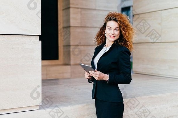 人、企业、现代科技理念。穿着黑色夹克和裙子的漂亮女商人站在办公室附近，手持现代平板电脑developi