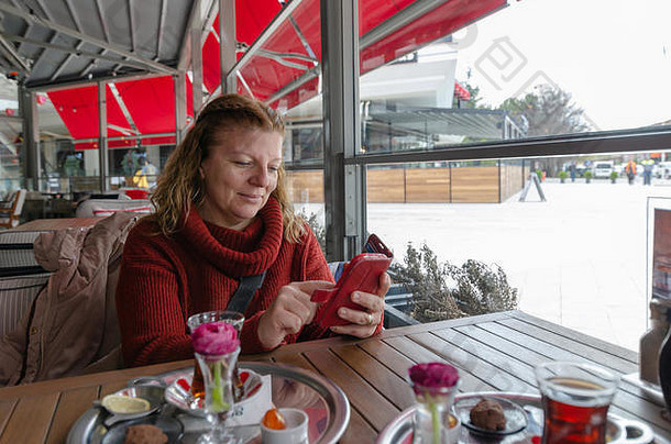 一位女士正在咖啡馆的智能手机上输入短信。一位金发的白人妇女坐在桌旁，用手机端着一杯茶。