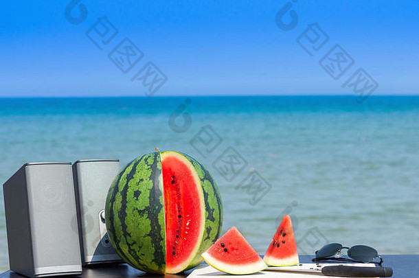 一对扬声器，整个西瓜和切好的西瓜，用小刀和木制砧板，还有一副太阳镜，在夏天的海滩上为夏日派对做准备。