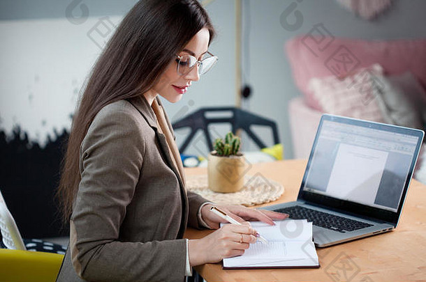 一个迷人的商业黑发女郎戴着眼镜计划一个商业<strong>项目</strong>，在<strong>计划表</strong>上做笔记。舒适时尚的办公室。
