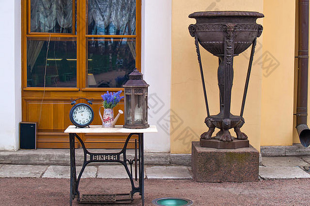 一个古老的机械闹钟，一个灯笼放在一个奇贡桌上的花瓶里，一个放在腿上的花瓶放在墙上，墙上有一个w形的大窗户