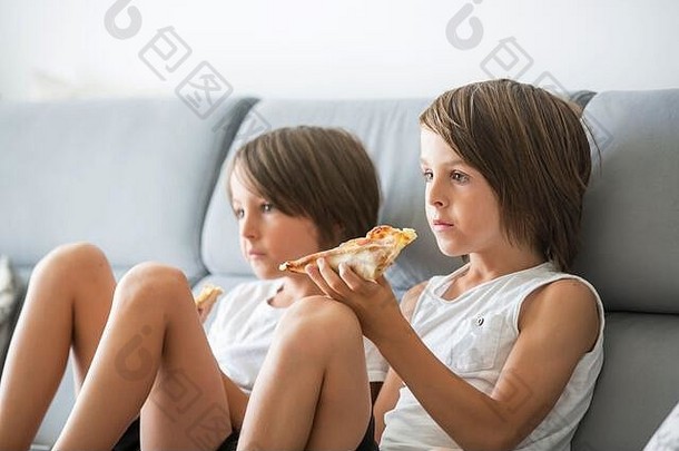 可爱的孩子，坐在沙发上，吃比萨饼，看电视。在家里的披萨派对上，饥饿的孩子在吃披萨