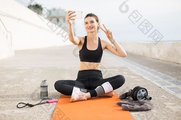 穿着运动上衣和紧身裤的年轻快乐女人坐在橙色瑜伽垫上，一边用两只手指在手机上愉快地拍照