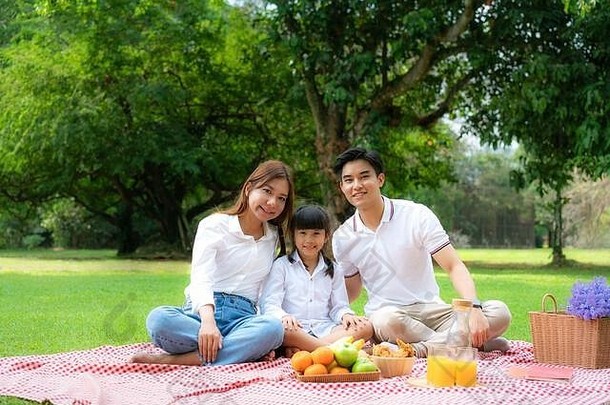 亚洲青少年家庭快乐假日野餐时刻在公园里，爸爸、妈妈和女儿看着相机，微笑着快乐地度过假期