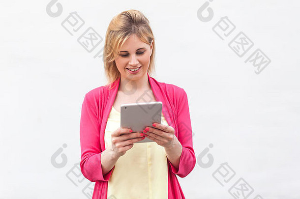 美丽成功的女商人的画像穿着粉红色上衣的年轻女子站着，用happy fac在她的平板电脑上使用和阅读有趣的信息