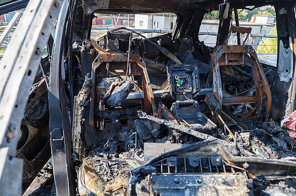 一辆在火灾或事故后被烧毁的汽车，停在一个布满铁锈和黑煤的停车场上，周围散落着零部件。抢劫、纵火、恐怖主义