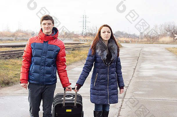 一对微笑的年轻夫妇提着一个手提箱，两人都握着提手，一起走在铁路沿线的一条乡村公路上。