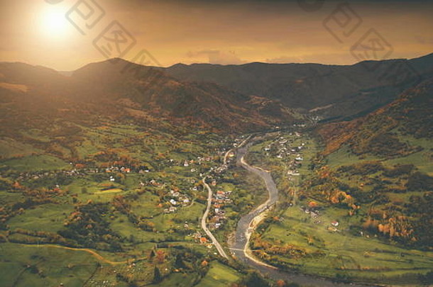 鸟瞰：美丽的峡谷村庄位于河边。壮观的秋山景观，绿草如茵，橘黄色的松林，日落多云的天空。喀尔巴阡山脉、乌克兰、欧洲。