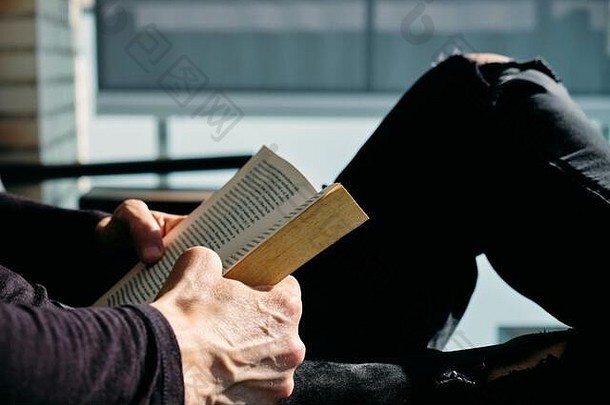 特写镜头：一个穿着休闲服的白人年轻人坐在阳台上的户外扶手椅上看书