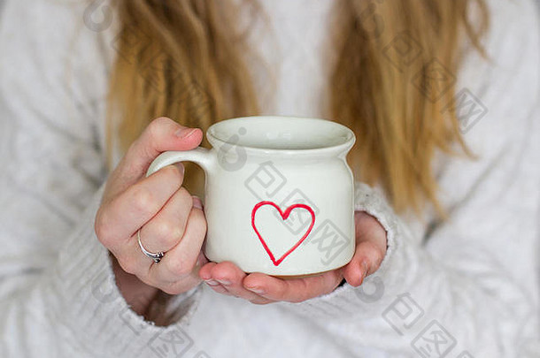 一个金发女孩的中间部分，手持一杯热乎乎的咖啡，带着一颗爱的心，穿着一件温暖的针织毛衣，展现了一种舒适的生活方式