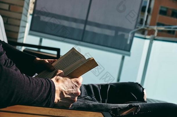 特写镜头：一个穿着休闲服的白人年轻人坐在阳台上的户外扶手椅上看书