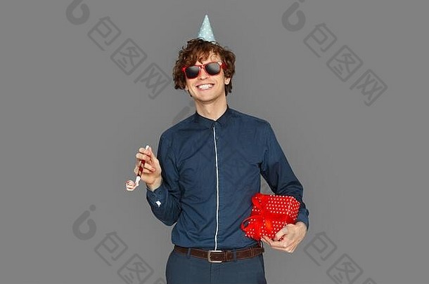 庆祝概念。戴着生日帽和太阳镜的少年男孩孤零零地站在灰色的地面上，手里拿着礼物和派对号角，脸上洋溢着喜悦的笑容