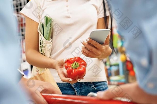日常购物。夫妇俩在超市里手推车一起拿着甜椒浏览智能手机特写镜头