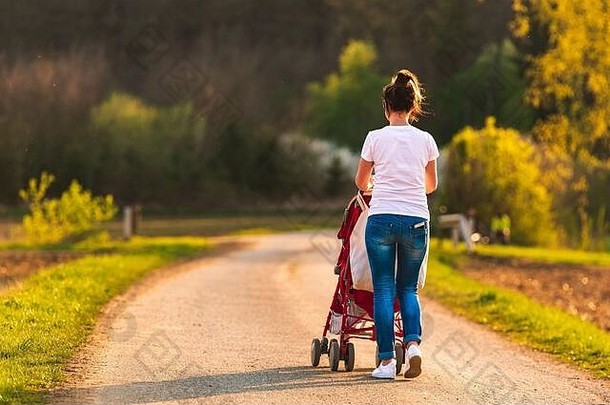 日落前，坐在红色婴儿车里的年轻母亲和孩子在农村户外散步。