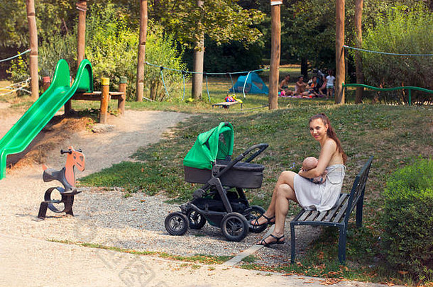 快乐的母亲在公共场所母乳喂养新生儿，婴儿车在户外行走时喂养婴儿，儿童游乐场和自然环境