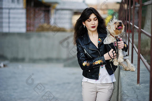 深色头发的吉普赛女孩和约克郡梗狗对着钢栏杆摆姿势。模特穿皮夹克和t恤，带装饰，裤子。