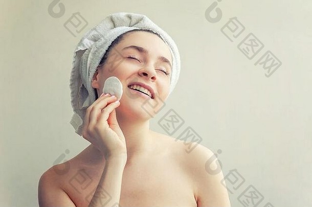 面带微笑的女人的美丽肖像，头戴毛巾，皮肤柔软健康，用白色背景上隔离的化妆棉<strong>卸妆</strong>。护肤洁面spa放松概念