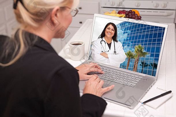 坐在厨房里的女人肩上使用笔记本电脑——在屏幕上与护士或医生在线聊天。