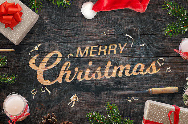雕刻在木制表面上的圣诞祝福语。圣诞礼物，树枝，凿子，蜡烛，松果和旁边的圣诞老人帽子。