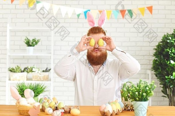 复活节快乐。一个滑稽的胖子坐在桌子旁装饰彩蛋，背景是复活节装饰