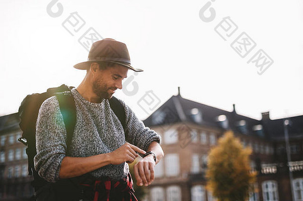 戴着旅游包和帽子的年轻人看着他的智能手表。游客微笑着看着他的手表，背景是阳光。