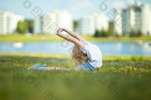 在一个阳光明媚的日子里，一个金发女孩在湖边绿草上的城市公园里跳舞、跳跃、做杂技和摆姿势