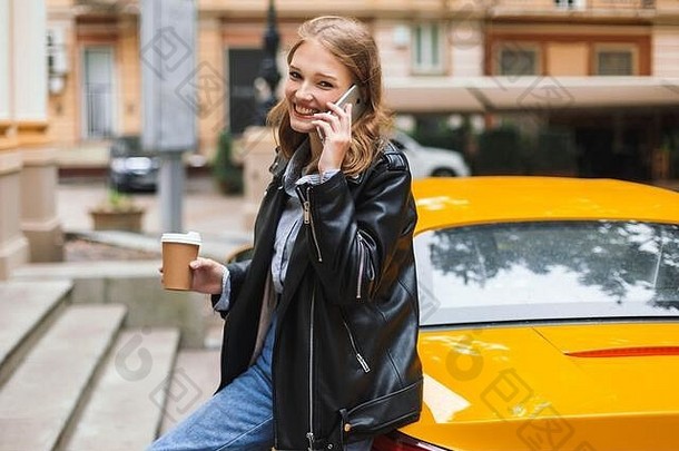 美丽的微笑女孩皮革夹克杯咖啡倾斜黄色的体育运动车会说话的手机幸福的相机