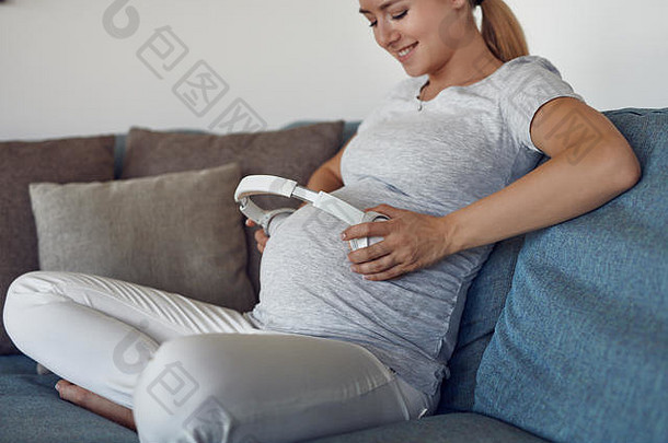 微笑快乐的孕妇坐着时将一套立体声耳机放在腹部，用耳机为未出生的婴儿播放音乐
