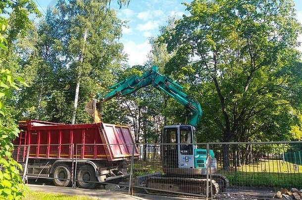 道路维修。在城市的庭院里，在金属栅栏后面的草地和树木中，一台绿松石挖掘机挖掘出一条路基。附近有一辆红色的自卸卡车。俄罗斯圣彼得堡市，2019年8月。