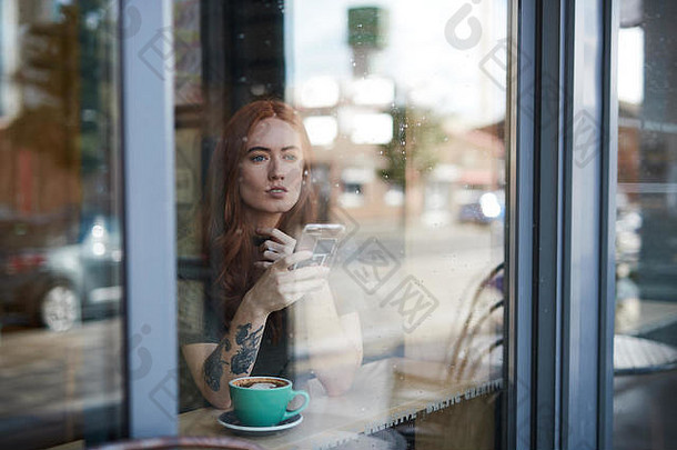 英国利物浦，一个手臂上有纹身的红头发女孩，独自坐在一家<strong>咖啡</strong>馆里，透过窗户看