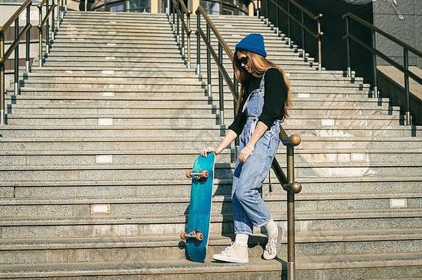 时髦穿着女人蓝色的牛仔布连身裤摆姿势滑板街照片肖像女孩持有滑板生活方式青年概念