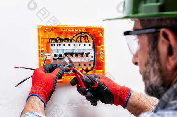 电工技术员戴着头盔、护目镜，双手戴着手套，用专业剪刀切割住宅配电盘的电缆