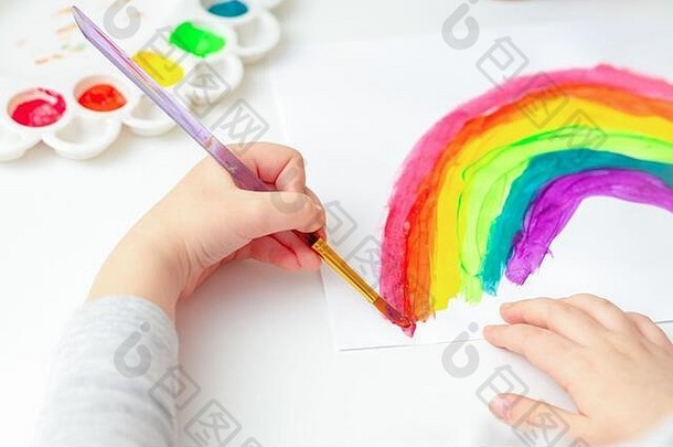 用画笔和颜料在白纸上画彩虹的孩子手的俯视图。