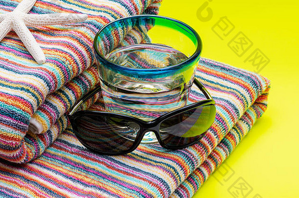 折叠式彩色条纹有机棉沙滩巾、蓝边水杯和黄色背景的黑色太阳镜是暑假的概念。