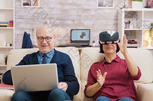 上了年纪的年龄女人坐着沙发穿虚拟现实护目镜男人。坐着沙发移动PC妻子