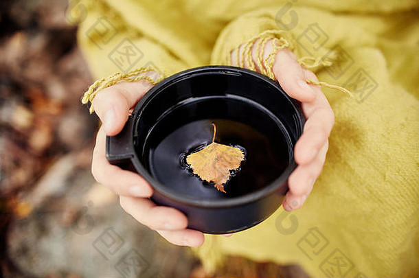 秋天，在金黄色的树叶覆盖的森林里，女人手里拿着一杯热茶。秋天来了，神奇的心情。茶叶中飘浮的黄叶