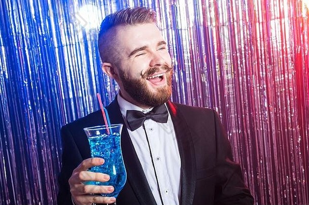 俱乐部派对和假日概念-优雅快乐帅哥的肖像，穿着昂贵的西装，在闪亮的背景上拿着蓝色鸡尾酒。