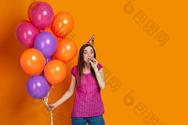 快乐有趣的年轻女子，穿着粉色t恤，戴着生日帽，橙色背景上有鲜艳多彩的气球吹着派对号角。文本空间