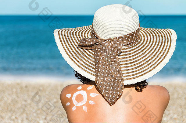 坐在海滩上戴着帽子的年轻女子肩上挂着防晒霜做成的太阳。