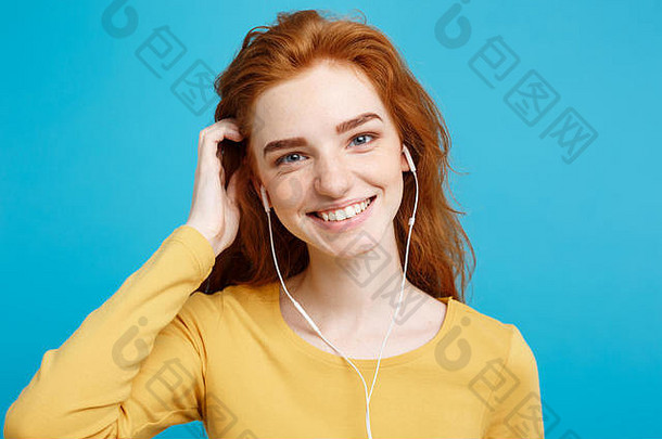 生活方式理念——欢快快乐的姜红色头发女孩的肖像，戴着耳机欣赏音乐，对着镜头欢快地微笑。在蓝色粉彩背景上隔离。空间。