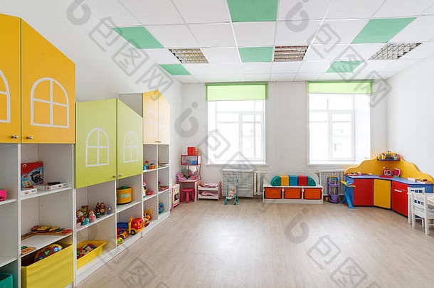 幼儿园宽敞的白色游戏室，有玩具、两扇大窗户和供上课用的桌子。
