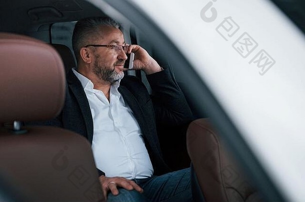 平静和良好的心情。坐在现代豪华轿车的后座上打商务电话。戴眼镜和公务服的高级男子